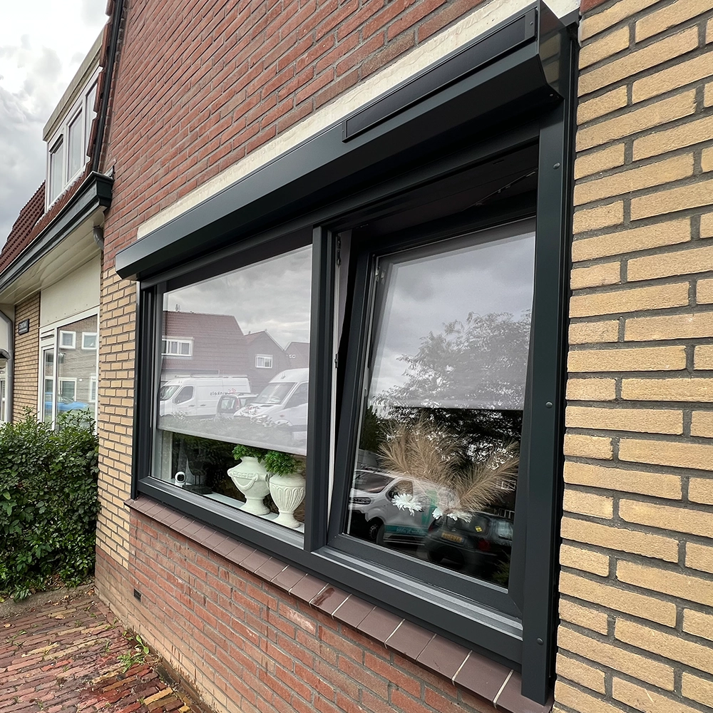 Donkergrijs kozijn met groot raam en kiepraam in een bakstenen gevel, met planten in de vensterbank.