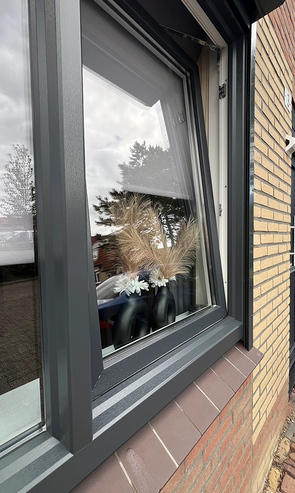 Donkergrijs kunststof kiepraam met vensterbank van bruine tegels in een bakstenen gevel.