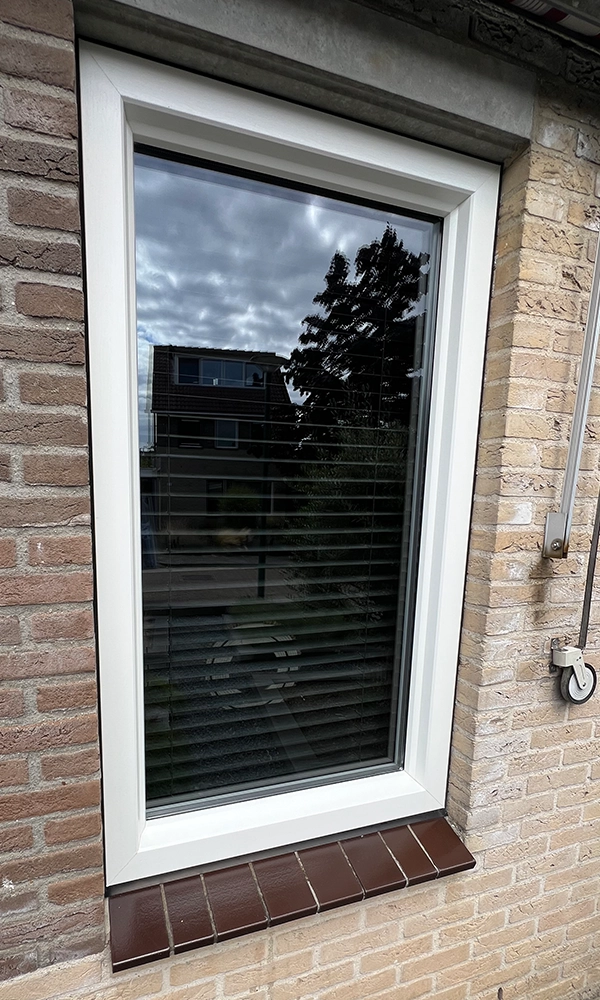 Klein raam met wit kunststof kozijn en bruine vensterbanktegels in een bakstenen gevel.