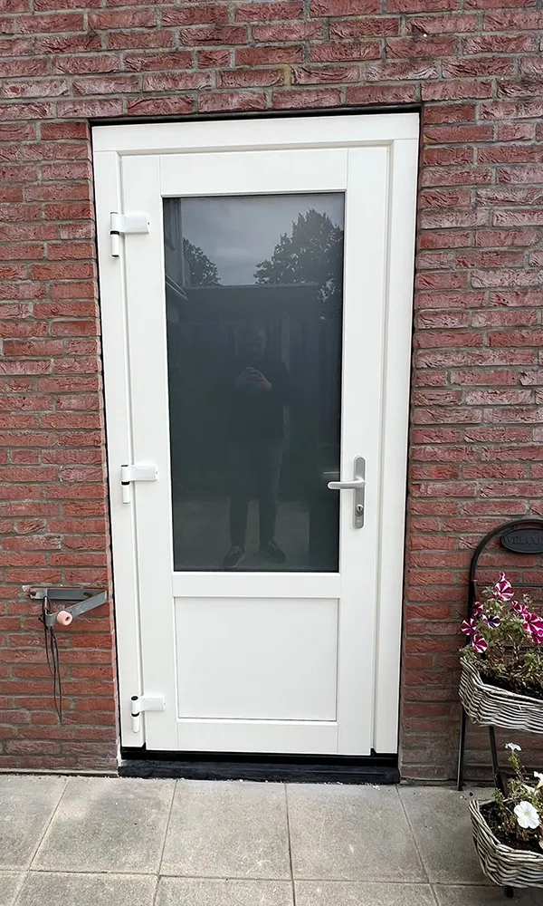 Witte kunststof deur met groot glasvlak in een rode bakstenen gevel, naast bloembakken met planten.