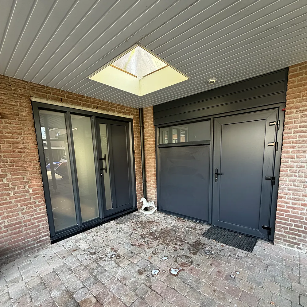 Antraciet kunststof achterdeur en schuifpui onder overkapping, geplaatst in bakstenen gevel met onderhoudsarme en strakke afwerking.