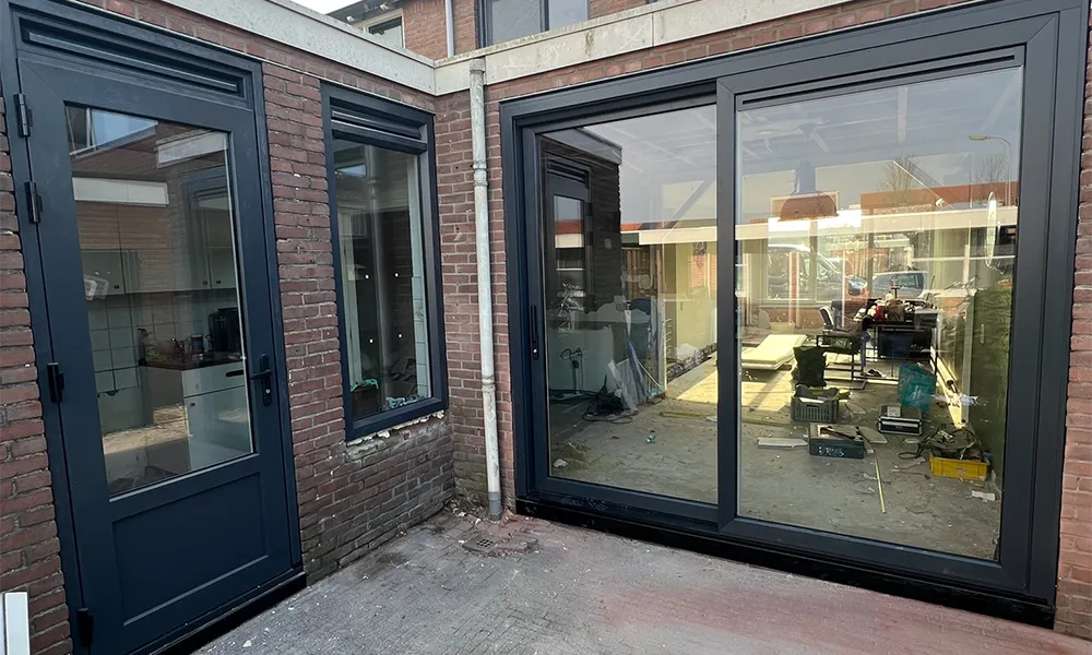 Antraciet kunststof schuifpui en achterdeur in bakstenen gevel, voorzien van grote glaspartijen en moderne, onderhoudsarme afwerking.