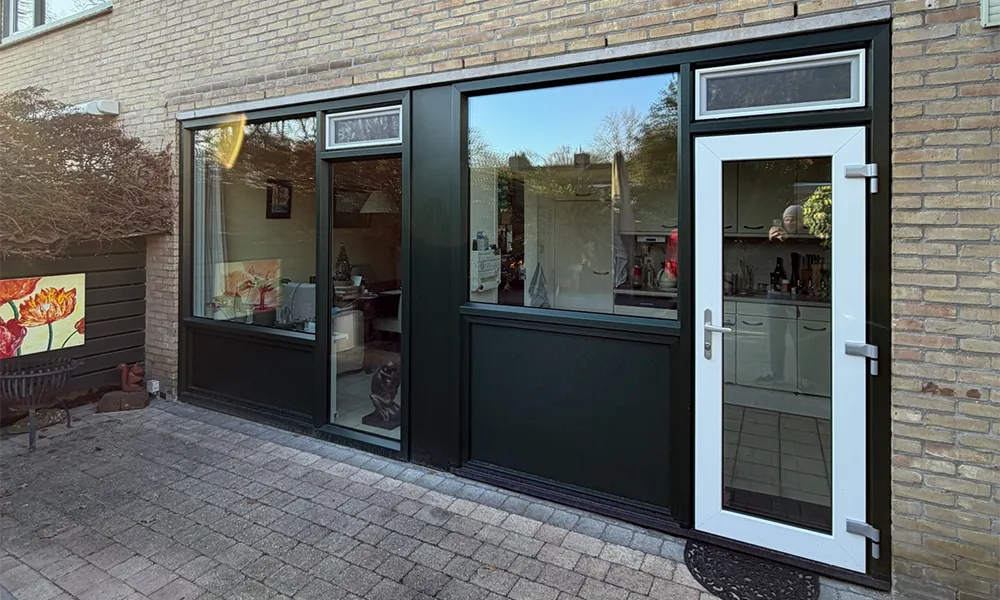 Donkergroene kunststof kozijnen en achterdeur met bovenlichten in bakstenen gevel, voorzien van grote glaspartijen en strakke afwerking.