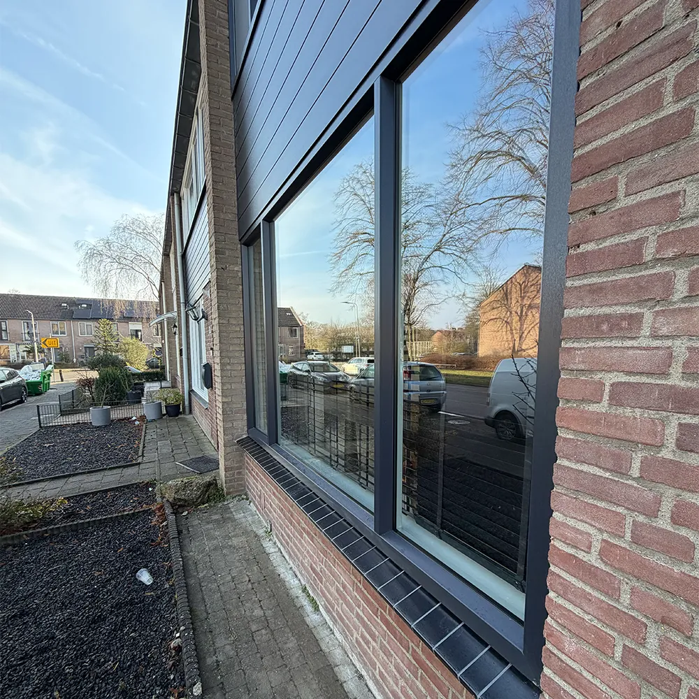 Detail van antracietkleurige kunststof kozijnen in bakstenen gevel met grote raampartij en strakke afwerking aan de buitenzijde.