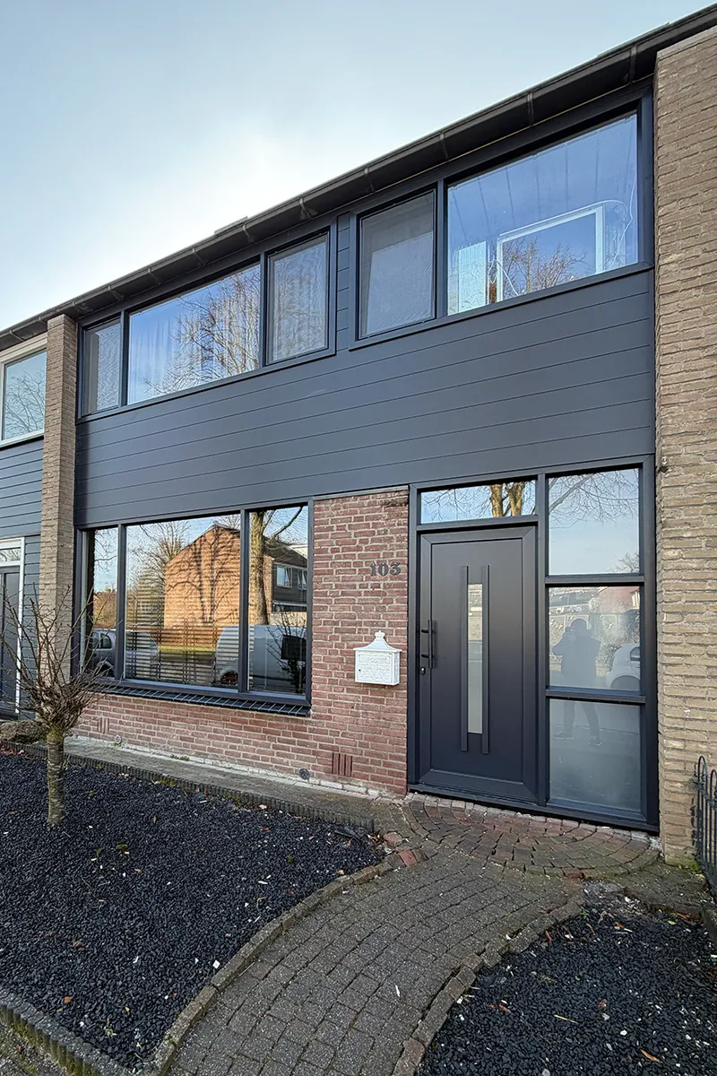 Moderne woning met antracietkleurige kunststof kozijnen, strakke voordeur en donkere gevelbekleding boven bakstenen gevel.