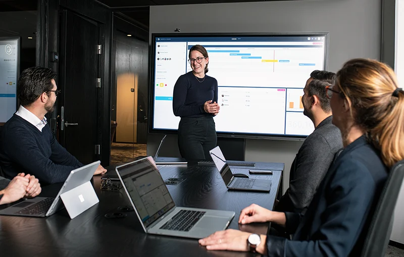 Business team in a meeting room reviewing a digital project management board on a large screen.