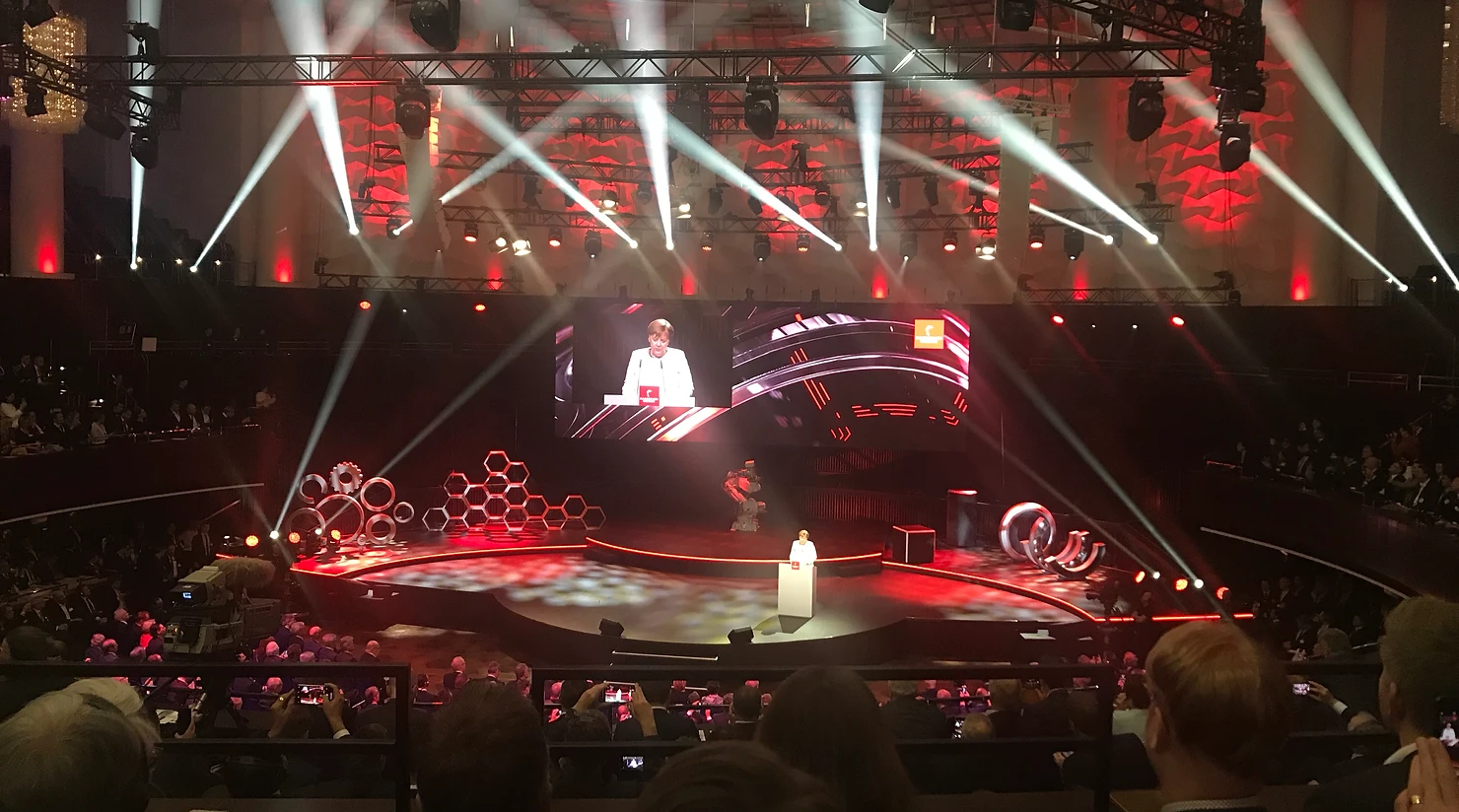 speaker presenting on stage at a large event with red lighting and spotlights.