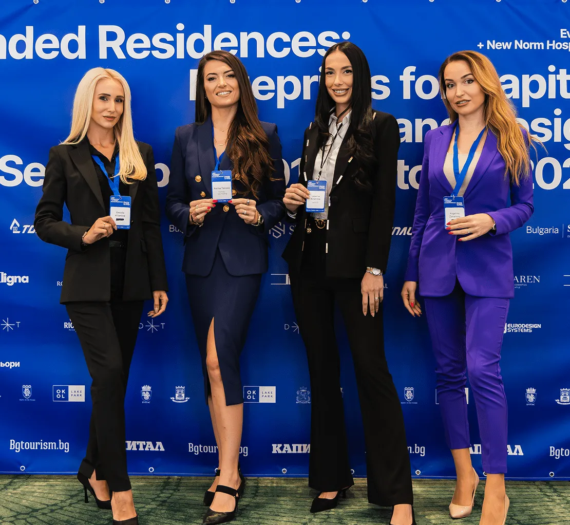 Четири професионалистки в бизнес костюми, стоящи пред син фон за конференция, държащи баджове с имена.