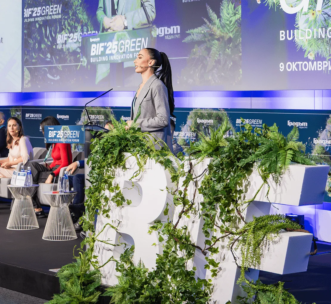 Жена в сиво сако говори на подиум, украсен със зеленина, на Форума за иновации в зеленото строителство BIF 25.