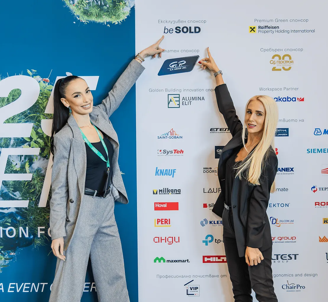 Две жени в професионално облекло се усмихват и сочат нагоре към логата на спонсорите на фона на конференция.