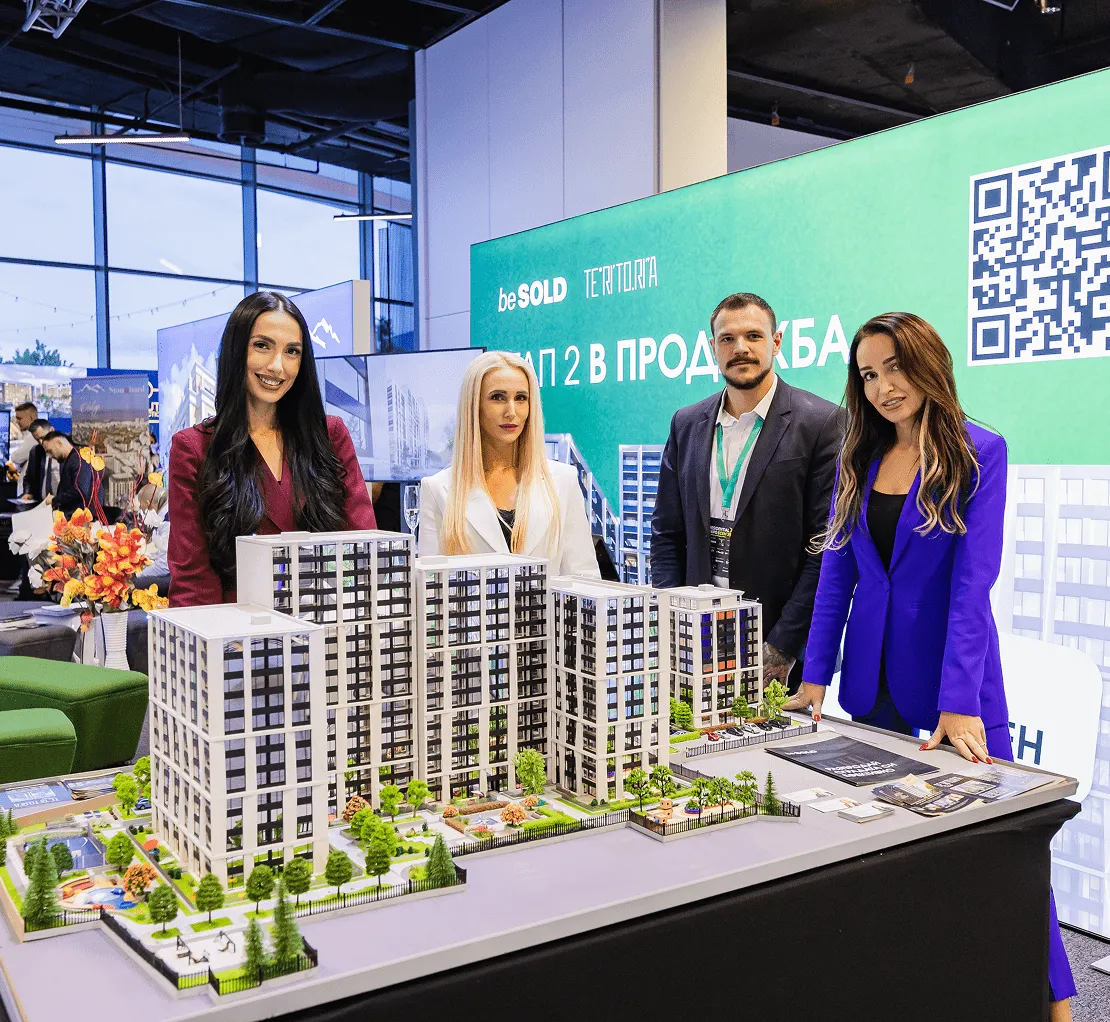 Четирима професионално облечени агенти за недвижими имоти позират зад подробен архитектурен модел на модерни жилищни сгради на щанд на търговско изложение.