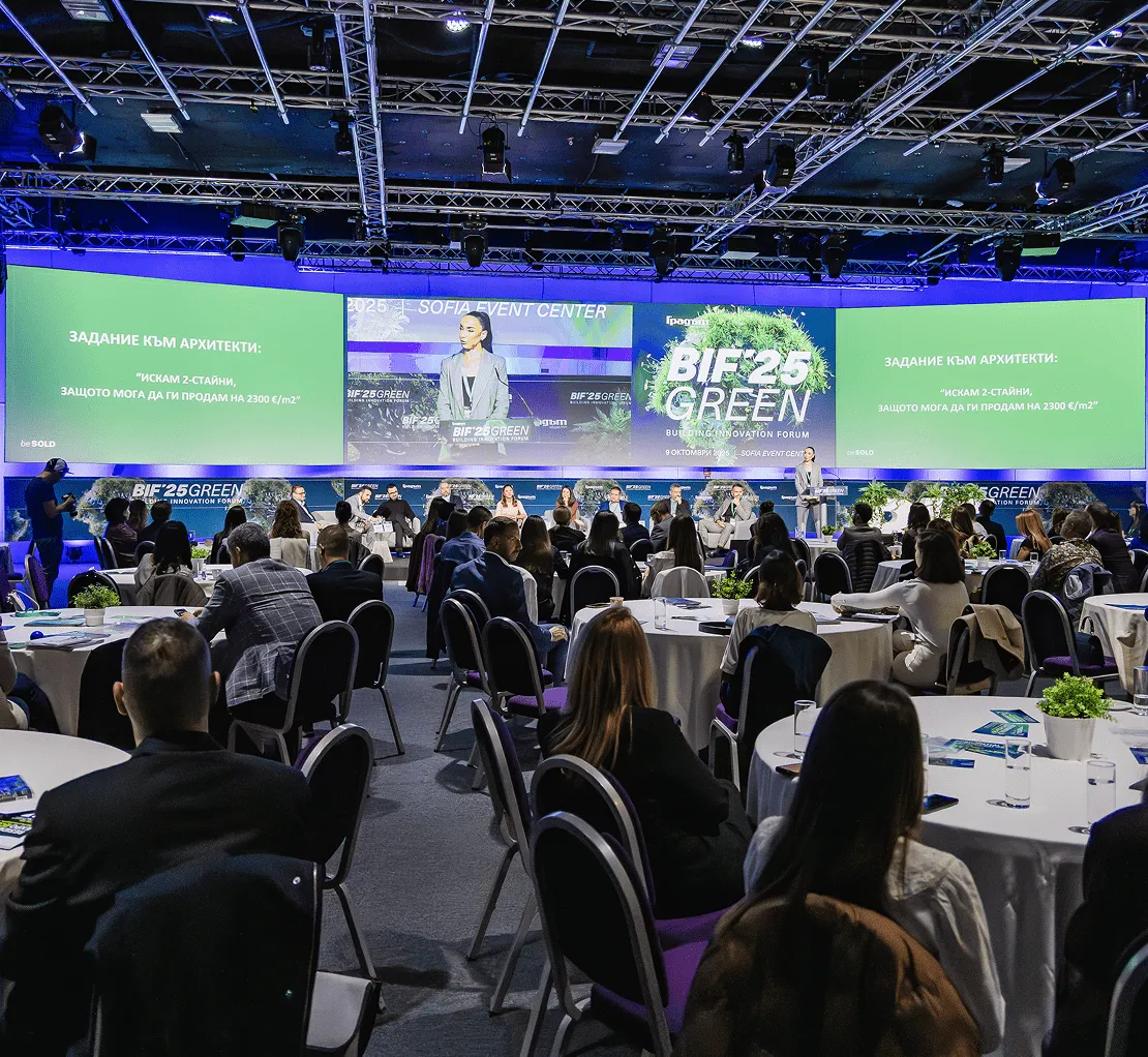 Публика, седнала на кръгли маси, слуша лектор, представящ се на сцената на Форума за иновации в зеленото строителство BIF 25 с големи екрани на зелена тематика зад тях.