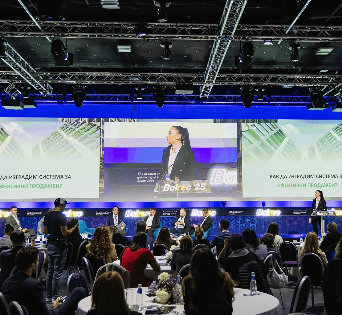 Сцена от конференция с жена лектор на подиум, участници в дискусията седнали на сцената и ангажирана публика, която наблюдава.