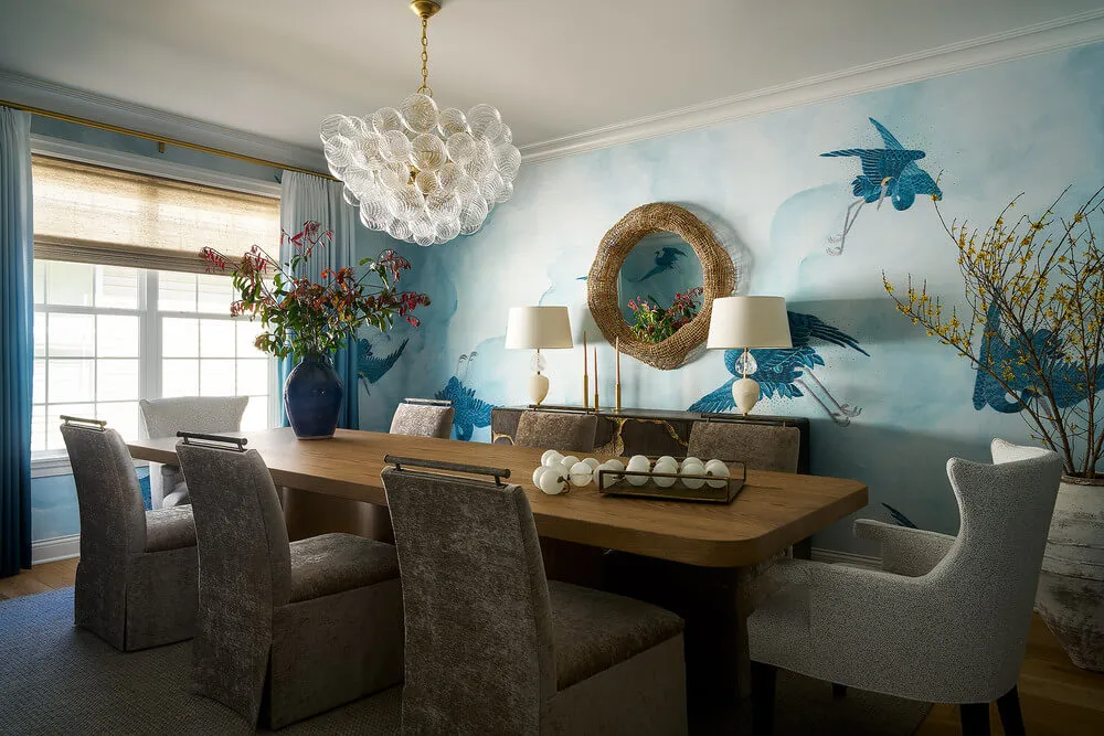 Dining room with a wooden table surrounded by gray upholstered chairs, a large blue vase with flowers, and a wall mural of blue birds under a glass bubble chandelier.