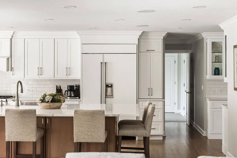 Modern kitchen with white cabinets, a large white refrigerator, marble countertop island, and four beige upholstered stools.