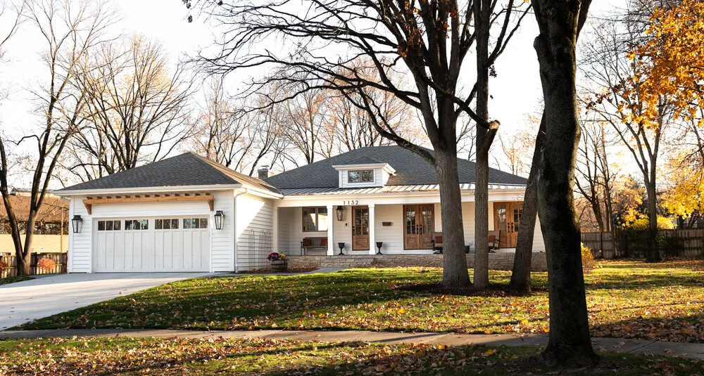 Single-story white house with a two-car garage, front porch, and large trees in a lawn with fallen autumn leaves.