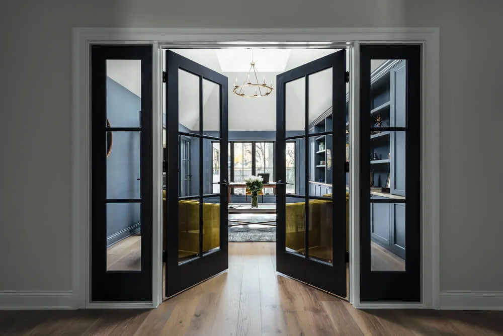 Open black-framed glass double doors leading to a modern home office with mustard velvet chairs, built-in shelves, and a chandelier.