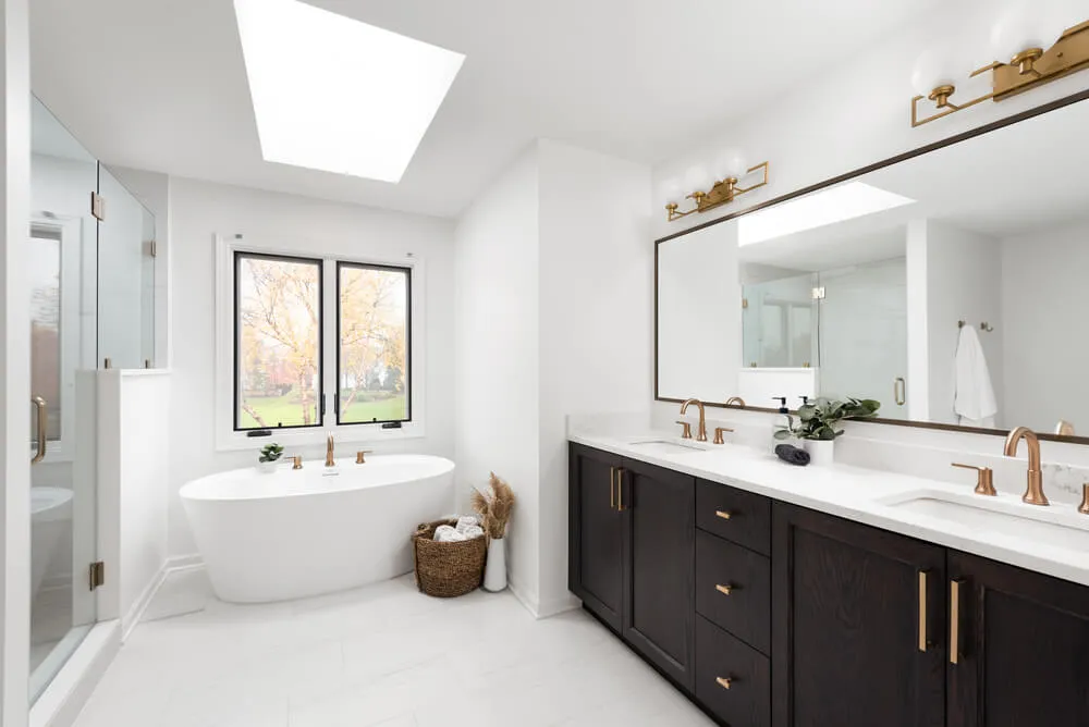 Modern bathroom with a freestanding white bathtub under a window, a large mirror above a double sink vanity with dark cabinets and gold fixtures.