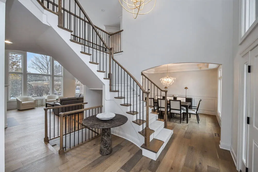 Modern foyer with curved staircase, round marble table, and views of a dining area and sitting room with large windows.
