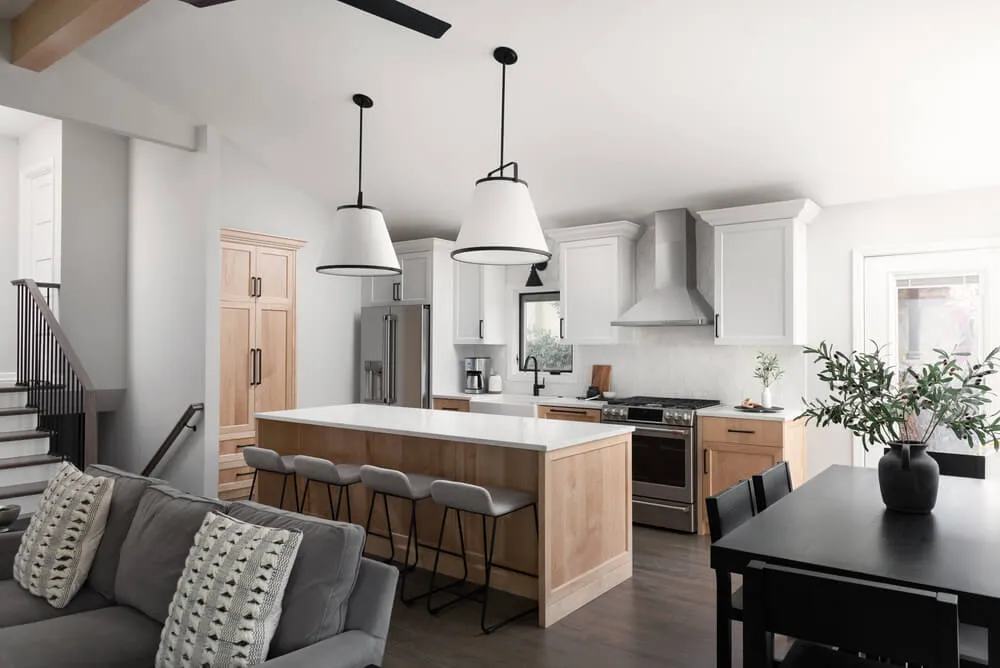Modern kitchen with a white island, four gray bar stools, pendant lights, wooden cabinets, stainless steel appliances, and a black dining table with a vase of greenery.