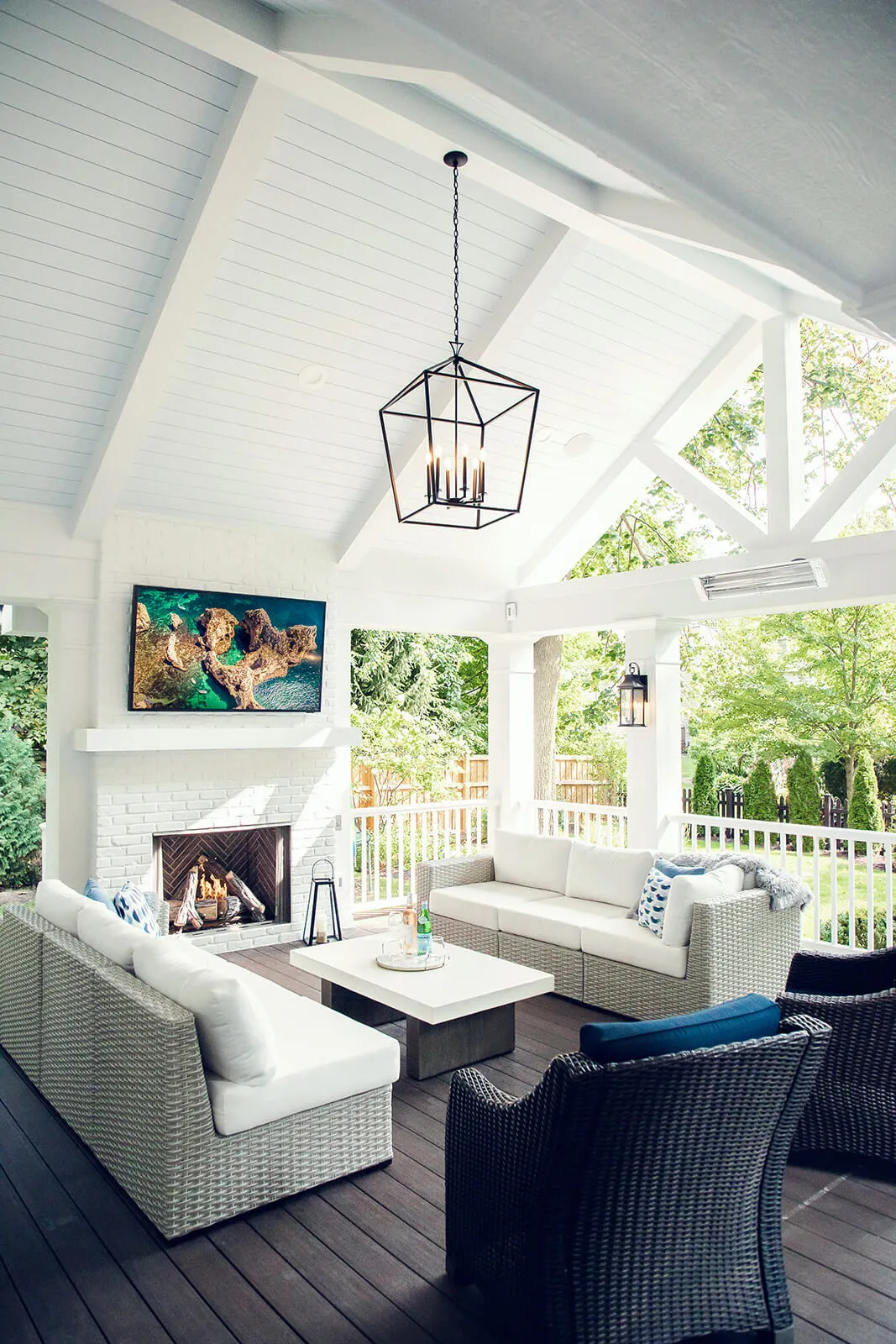 Covered outdoor patio with white cushioned wicker sofas, black wicker chairs, a white coffee table, a lit fireplace, and a TV mounted above it.