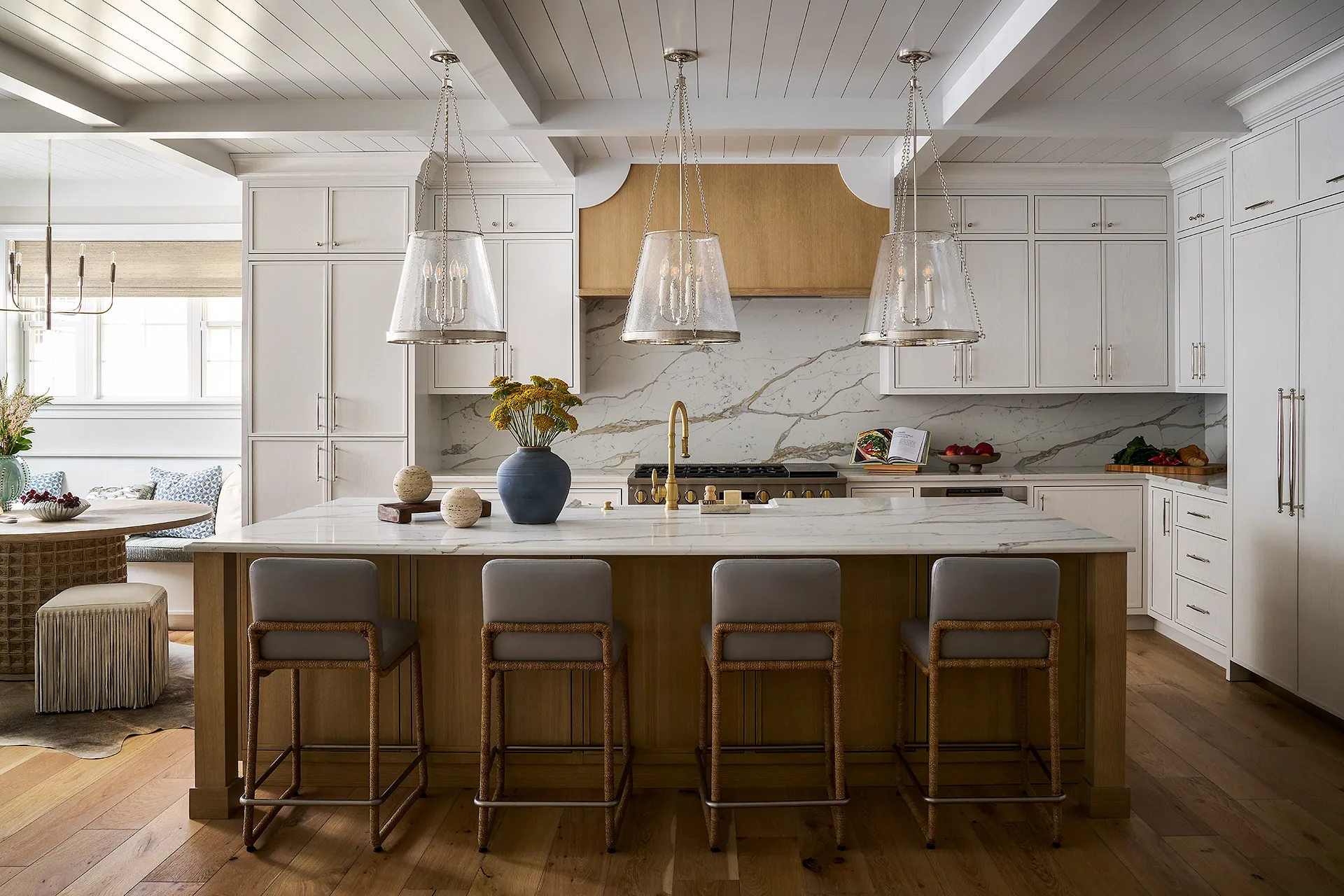 Modern kitchen with large marble island, four gray bar stools, three glass pendant lights, white cabinets, and wood flooring.