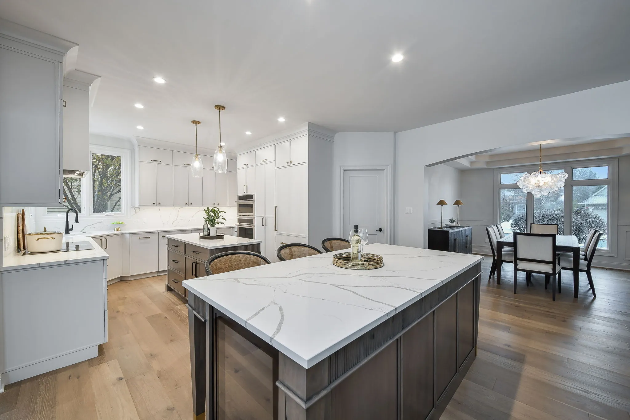 Spacious modern kitchen with white cabinetry, two large marble-topped islands, wicker bar stools, and a view into a dining area with large windows and a chandelier.