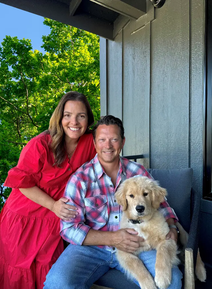 Smiling couple on a porch with green trees in the background; man holds a golden retriever puppy on his lap.