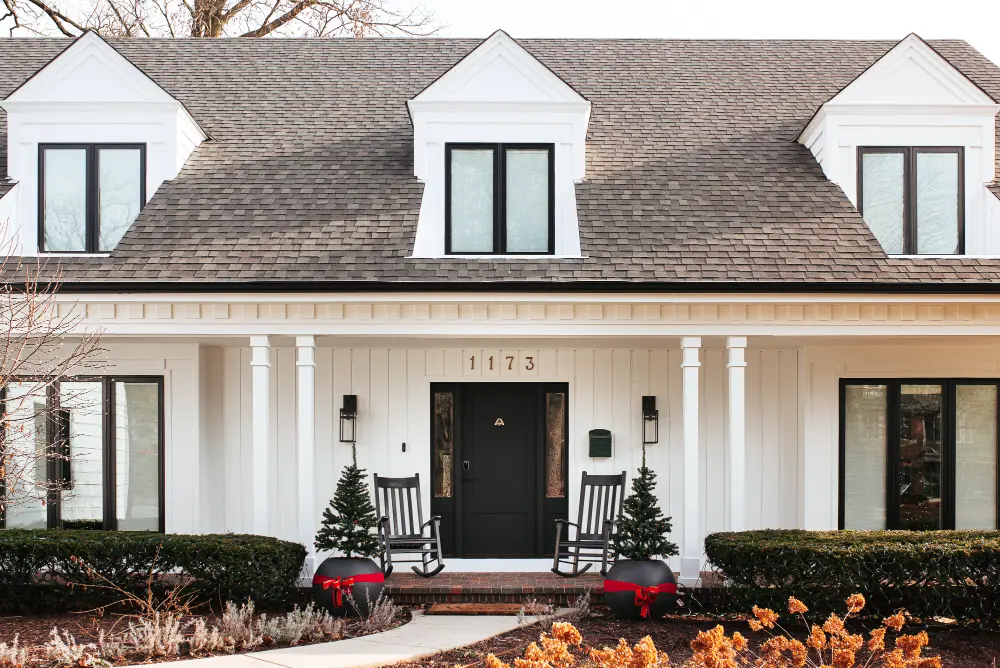 White house exterior with a black front door, two rocking chairs, small decorated evergreen trees in black pots with red bows, and three dormer windows on the roof.