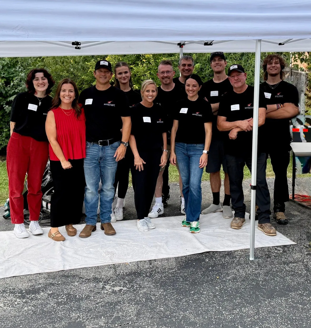 Group of eleven people smiling under a white canopy, most wearing black Redstart shirts, standing on a white cloth outdoors with greenery in the background.