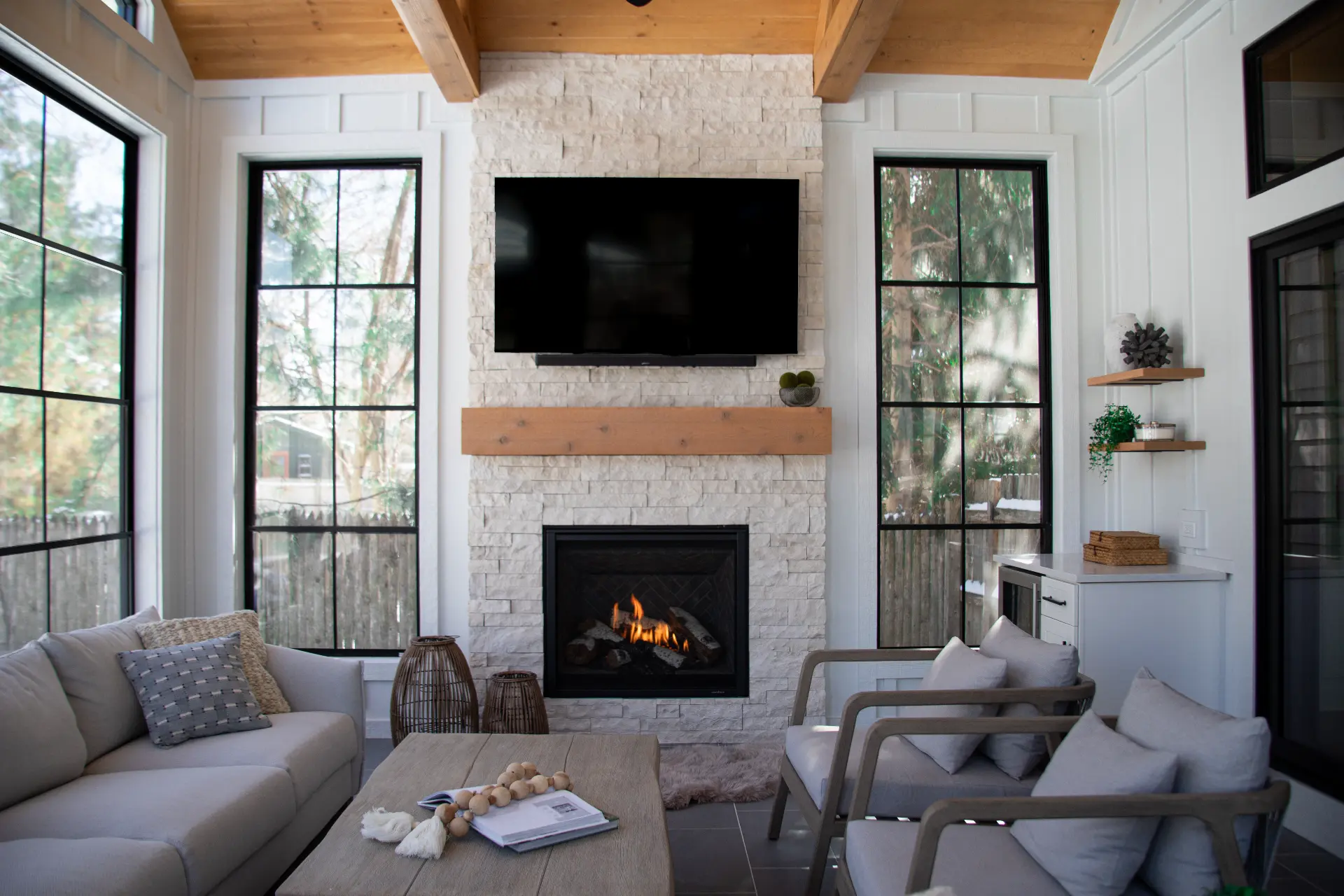 Cozy modern living room with a stone fireplace, mounted TV, large windows, beige sofa, and gray armchairs.