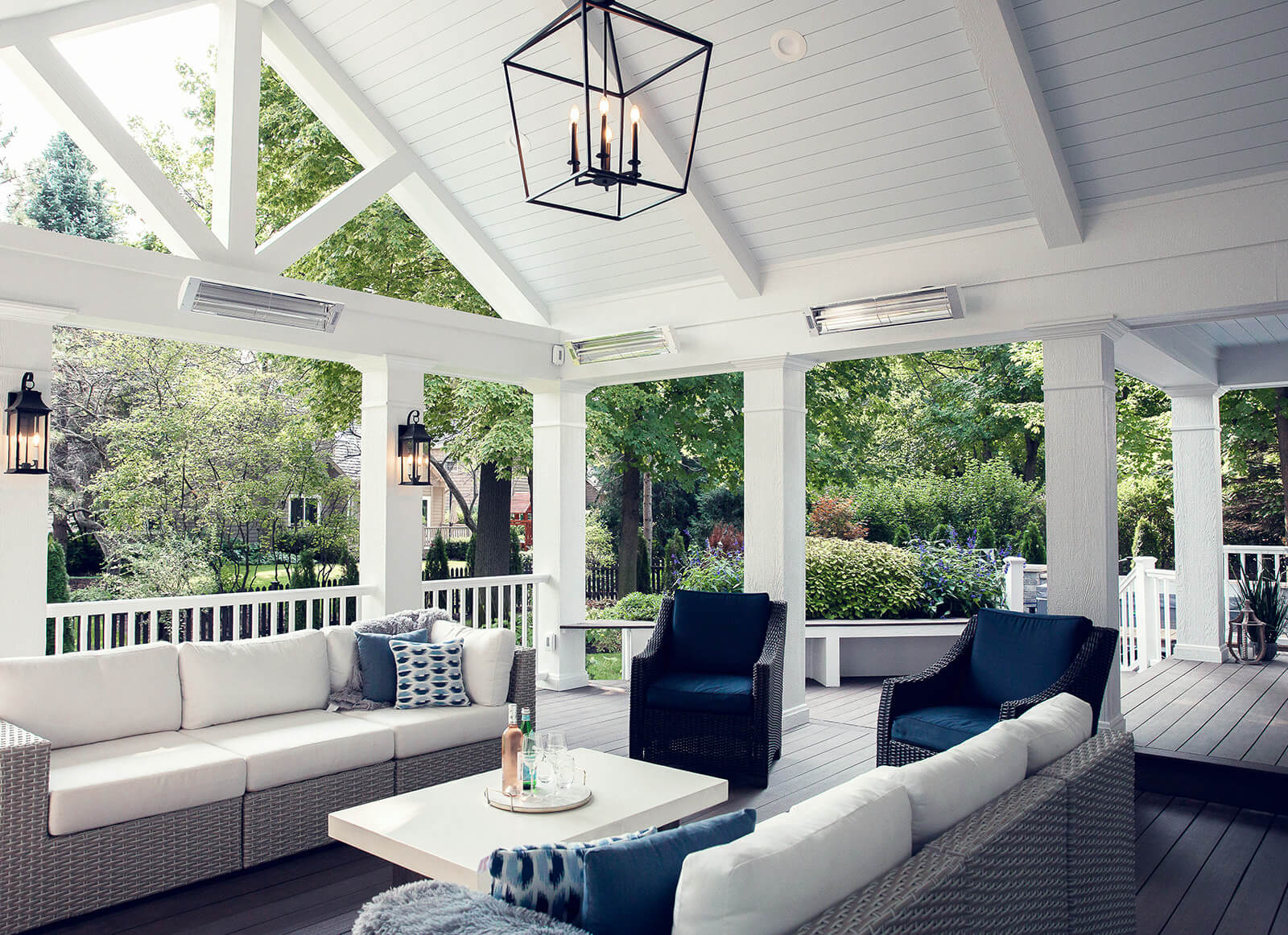 Covered outdoor porch with white cushioned wicker sofas, two dark blue cushioned wicker chairs, and a white coffee table with drinks, surrounded by green trees.