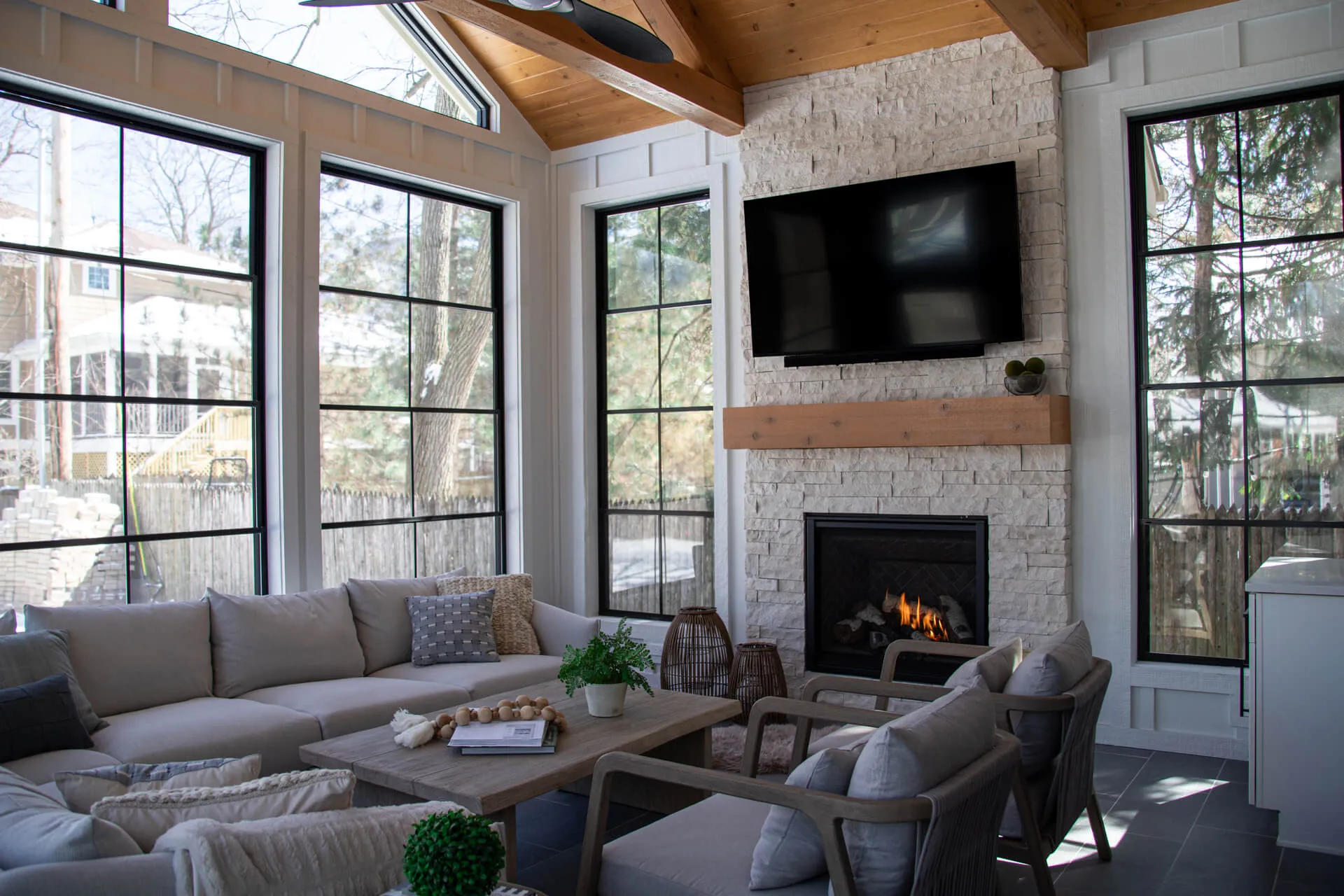 Bright living room with large windows, a stone fireplace with mounted TV, beige sectional sofa, and wooden coffee table.