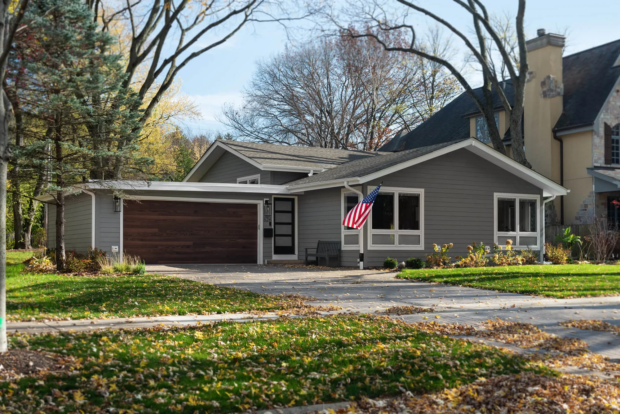 Single-story gray house with a wooden garage door, an American flag, and fallen autumn leaves on the lawn and driveway.
