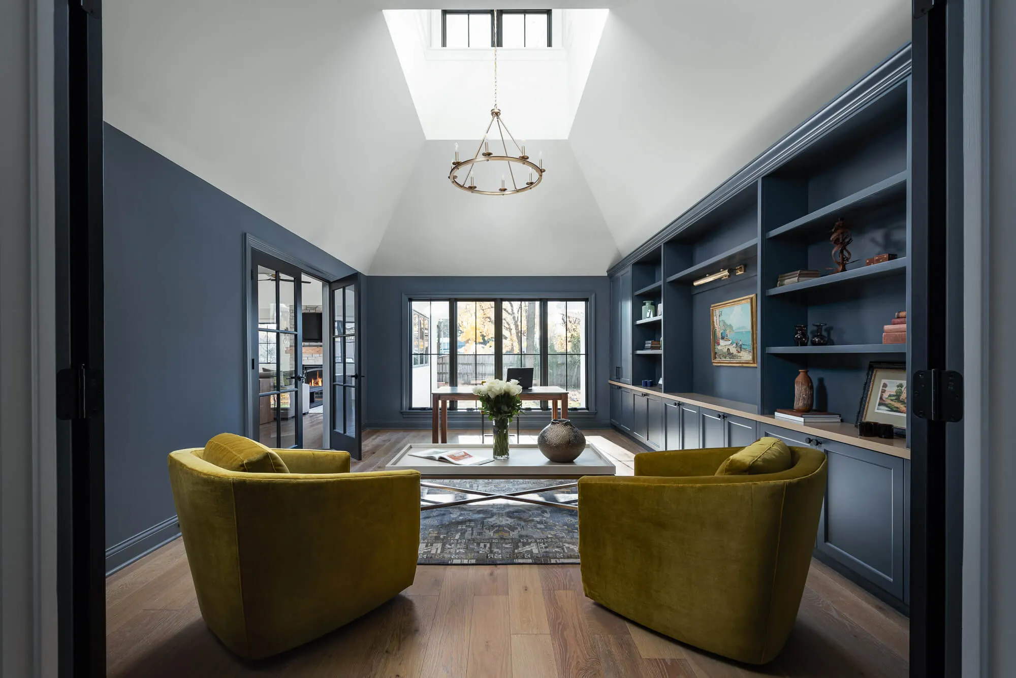 Modern home office with navy blue built-in shelves, large windows, two mustard yellow velvet armchairs, white coffee table with vase of flowers, and skylight ceiling.