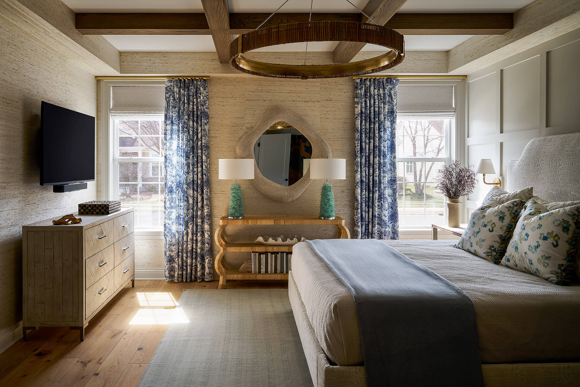Well-lit bedroom featuring a large bed with blue and floral pillows, a wooden dresser with a TV mounted above, blue patterned curtains, and a decorative round mirror above a wooden console with two green lamps.