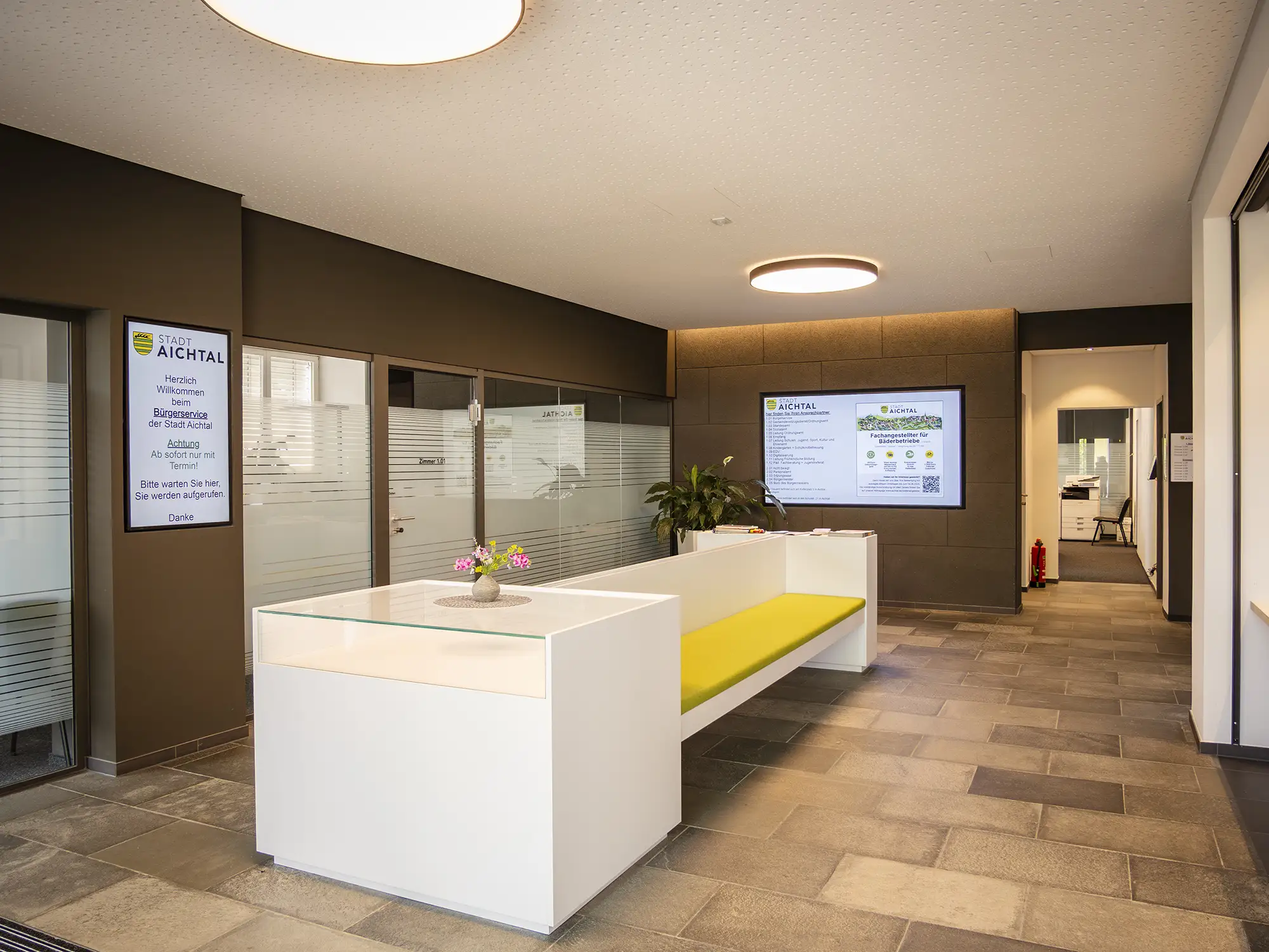 Moderne Empfangswartebereich mit weißer Theke, gelber Sitzbank, digitalen Informationsbildschirmen und Pflanze in einem Bürogebäude.
