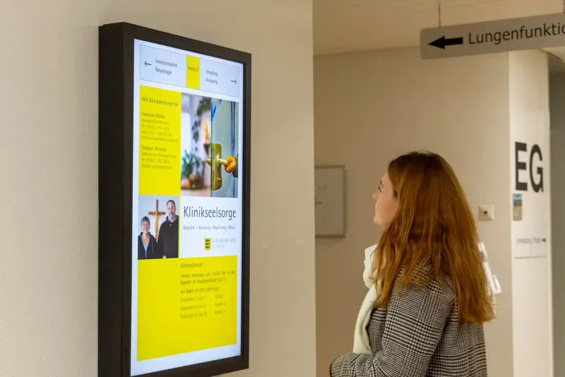 Eine Frau die auf ein digitales Infoboard in einem Klinikum schaut.