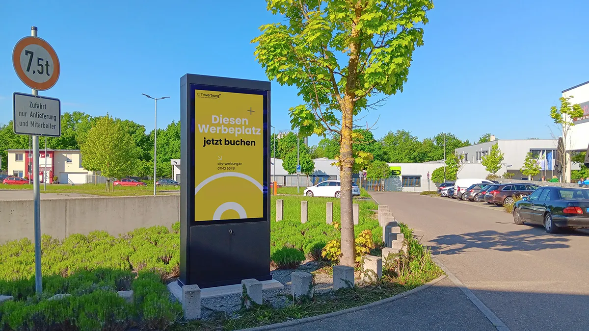 Werbestele mit gelbem Schild und der Aufschrift 'Diesen Werbeplatz jetzt buchen' neben Straße und Baum bei sonnigem Himmel.