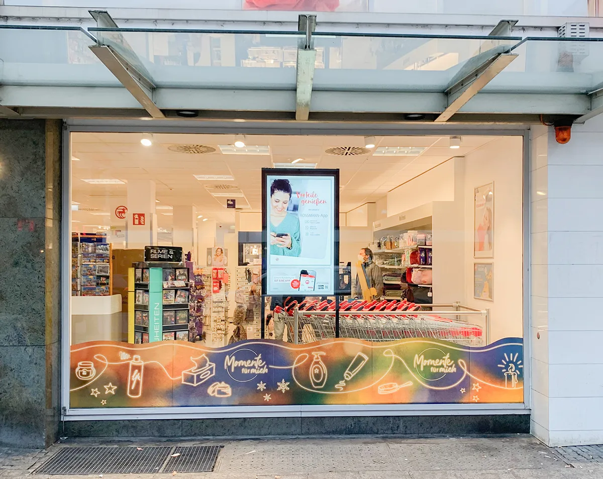 Schaufenster eines Rossmann-Drogeriemarkts mit Einkaufswagen und Werbeplakat innen.