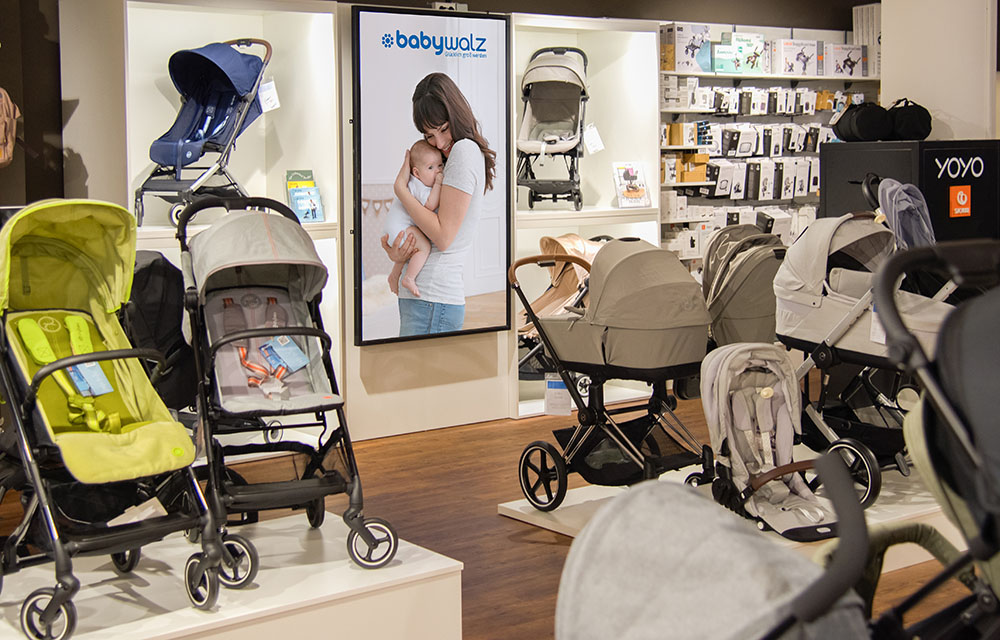 Mehrere Kinderwagen in verschiedenen Farben und Modellen in einem Babyausstattungsgeschäft mit einem Poster einer Frau, die ein Baby hält.
