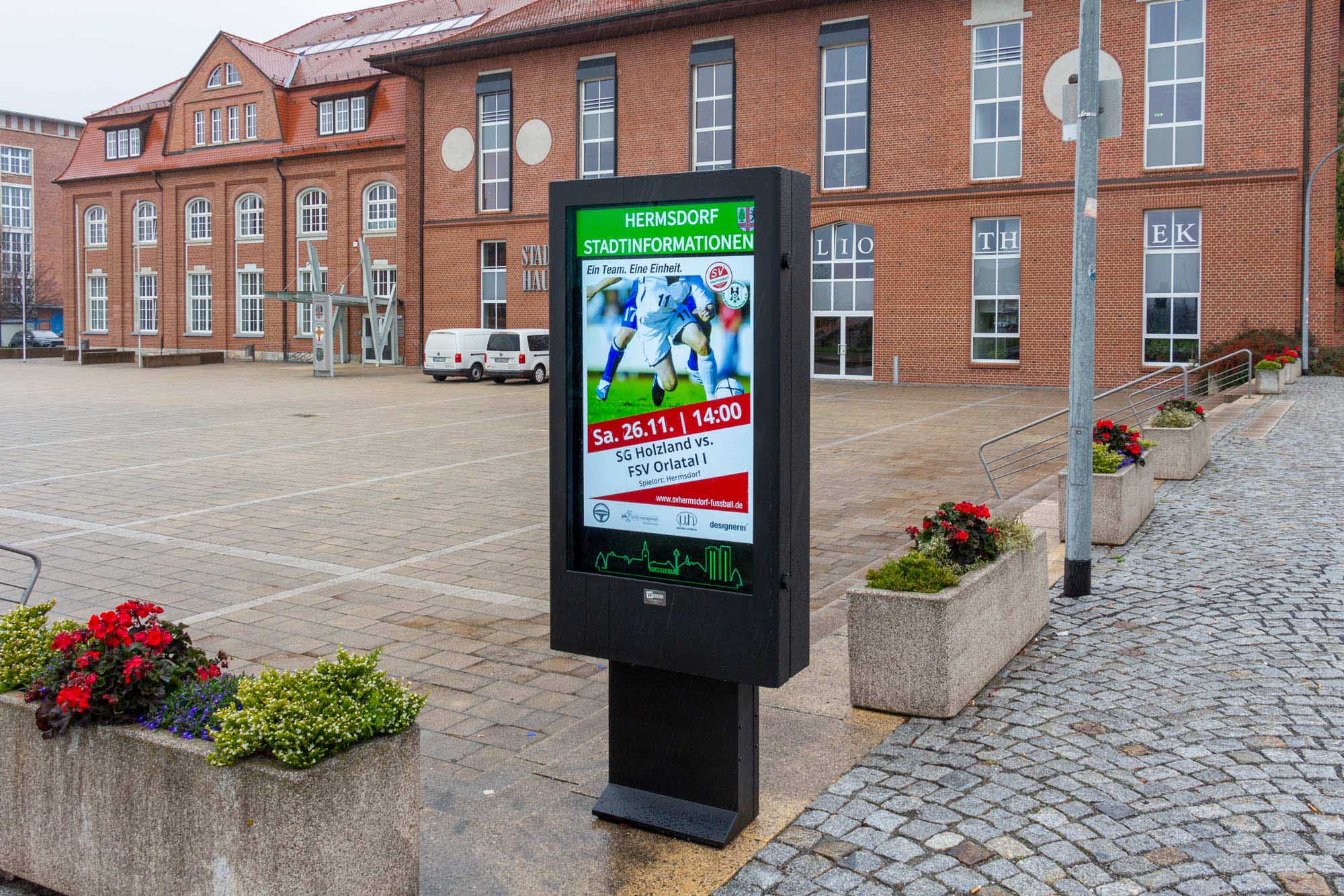 Digitale Informationsanzeige vor Rathaus in Hermsdorf zeigt Ankündigung eines Fußballspiels zwischen SG Holzland und FSV Orlatal.