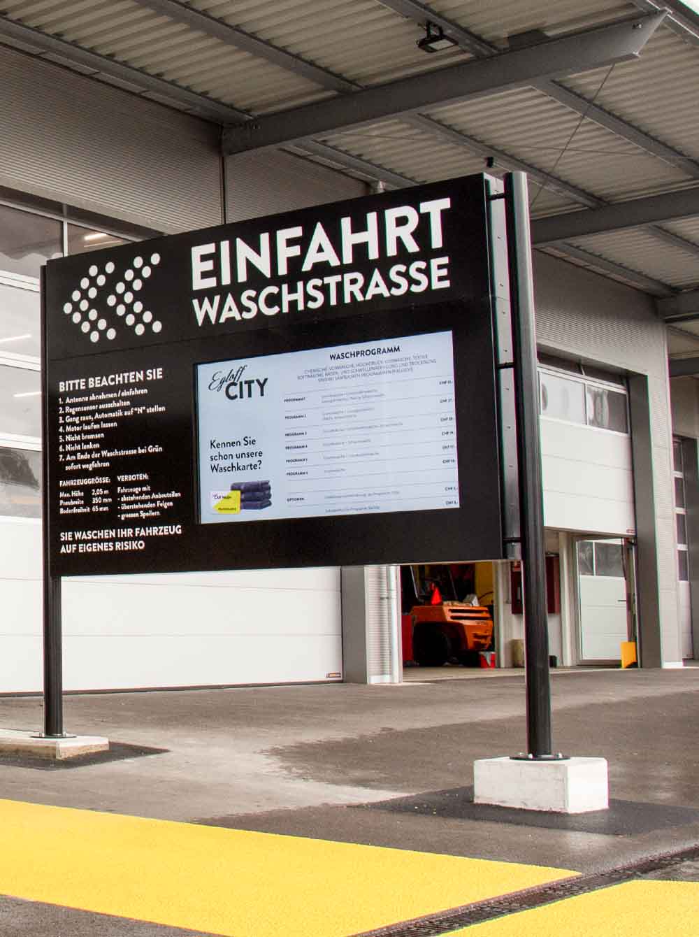 Digitales Schild mit weißer Aufschrift 'Einfahrt Waschstraße' vor einer Autowaschhalle mit geöffneter Toröffnung.