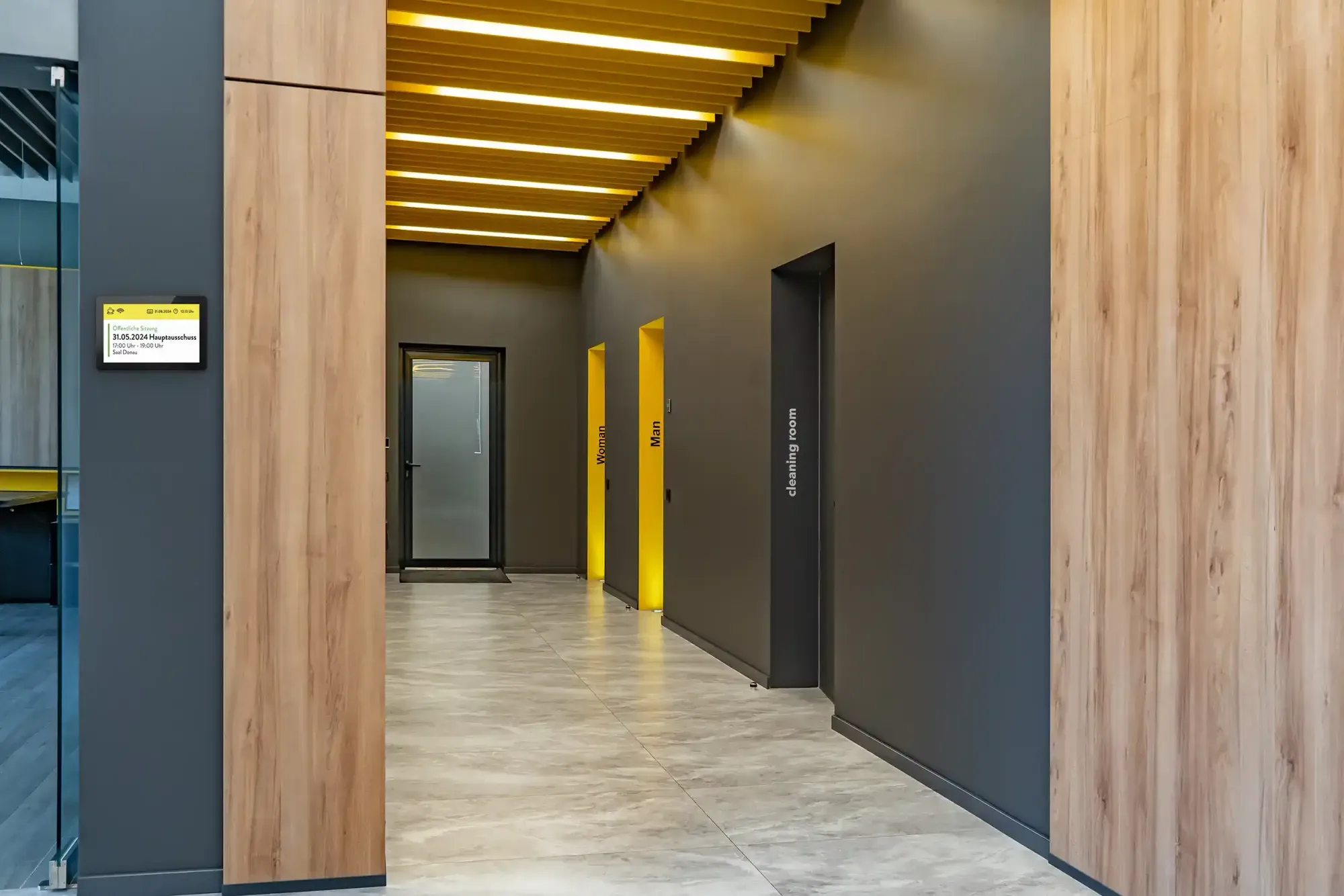 Moderner Flur mit grauen Wänden, gelben Türrahmen für Damen- und Herrentoiletten und Türschild mit Informationen an der Wand.