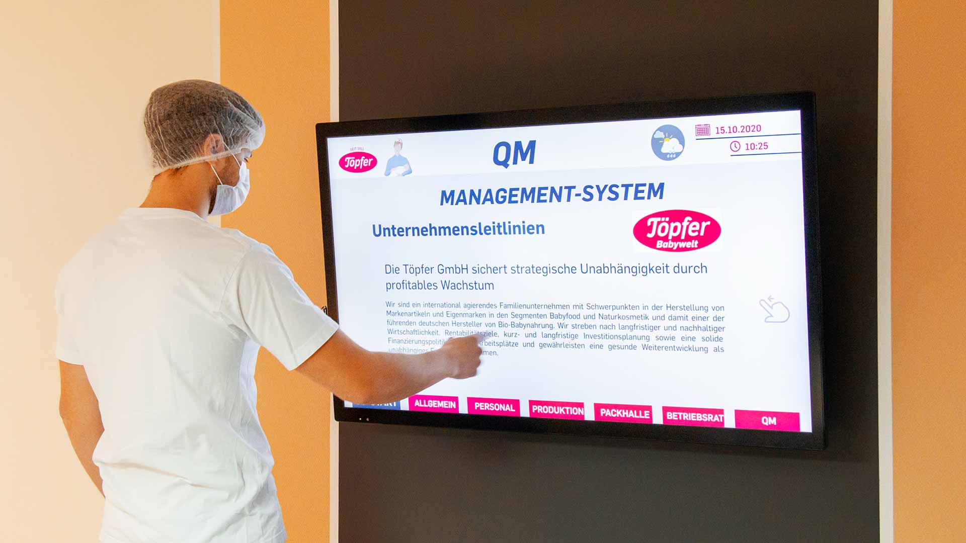 Mitarbeiter in weißem T-Shirt, Haarnetz und Maske zeigt auf digitalen Bildschirm mit Unternehmensleitlinien der Töpfer GmbH im QM-Management-System.