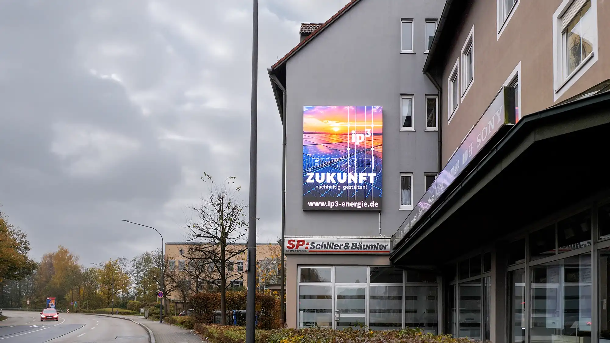 Digitale Werbetafel an der Seitenwand eines Gebäudes in einer Straßenumgebung mit bewölktem Himmel.