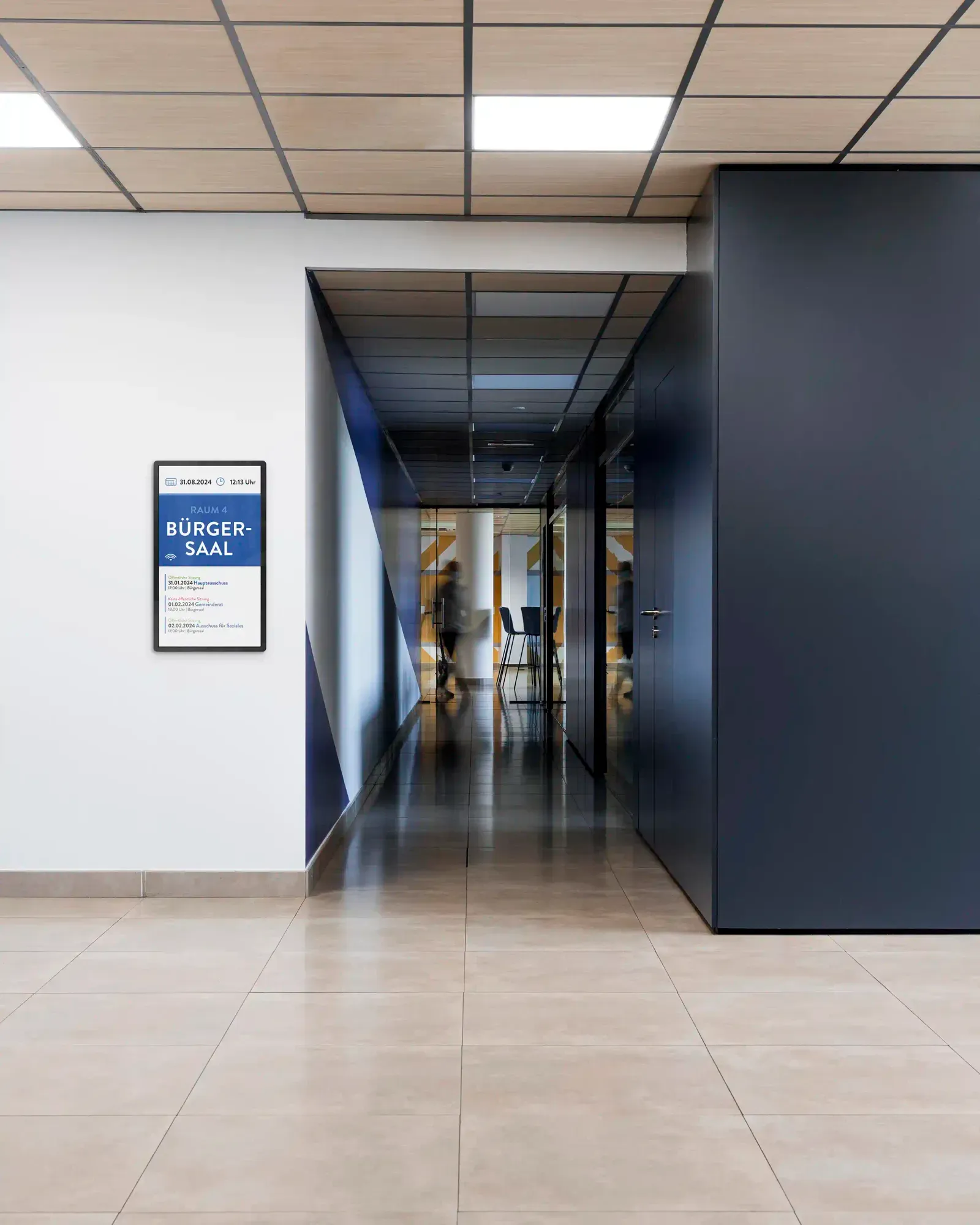 Moderner Büroflur mit Fliesengang, digitalem Türschild für Bürgersaal Raum 4 und verschwommener Person im Hintergrund.