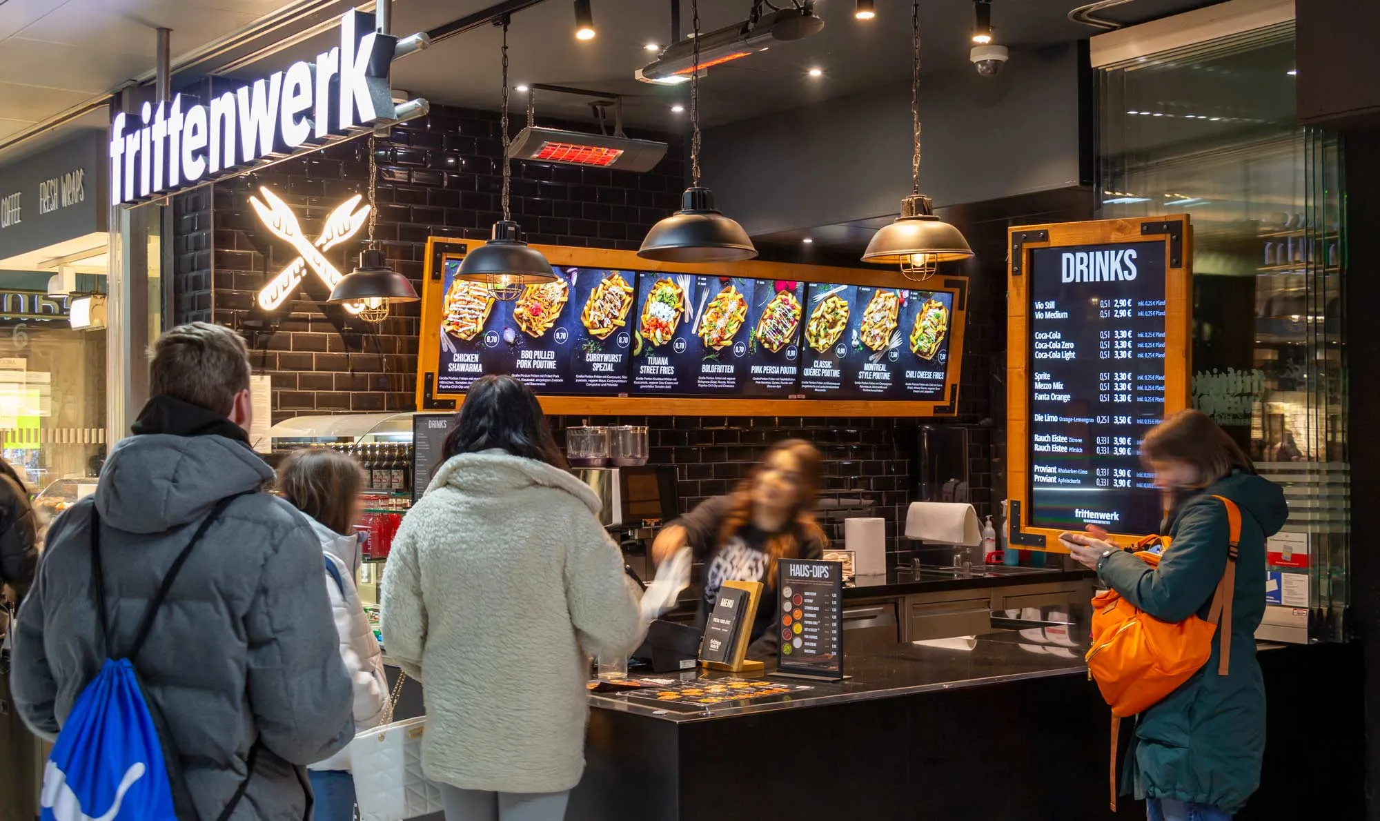 Menschen stehen an der Theke eines Frittenwerk Imbisses mit beleuchtetem Menü und Getränketafel in einer modernen Umgebung.