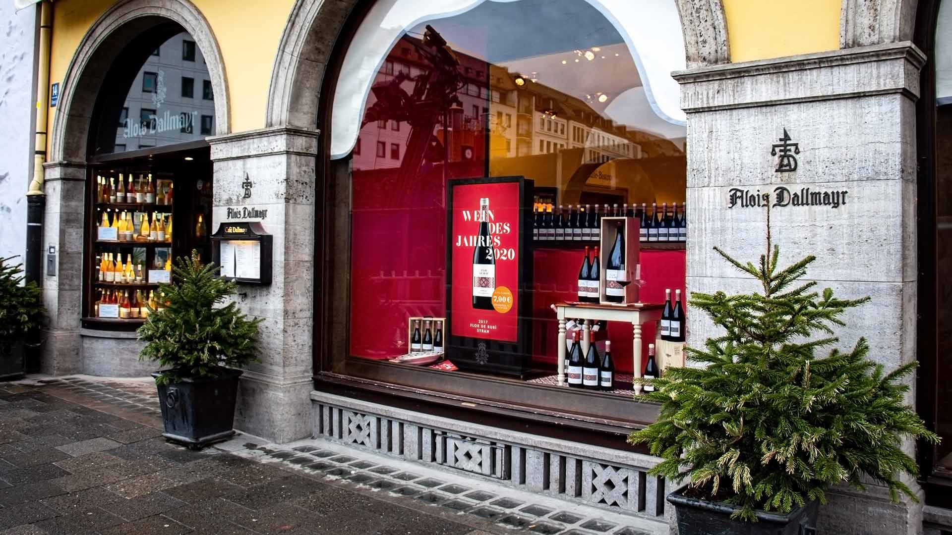 Außenansicht des Alois Dallmayr Ladens mit Weinflaschen im Schaufenster und Tannenbäumen davor. Im Fenster ein Schaufenster-Display.