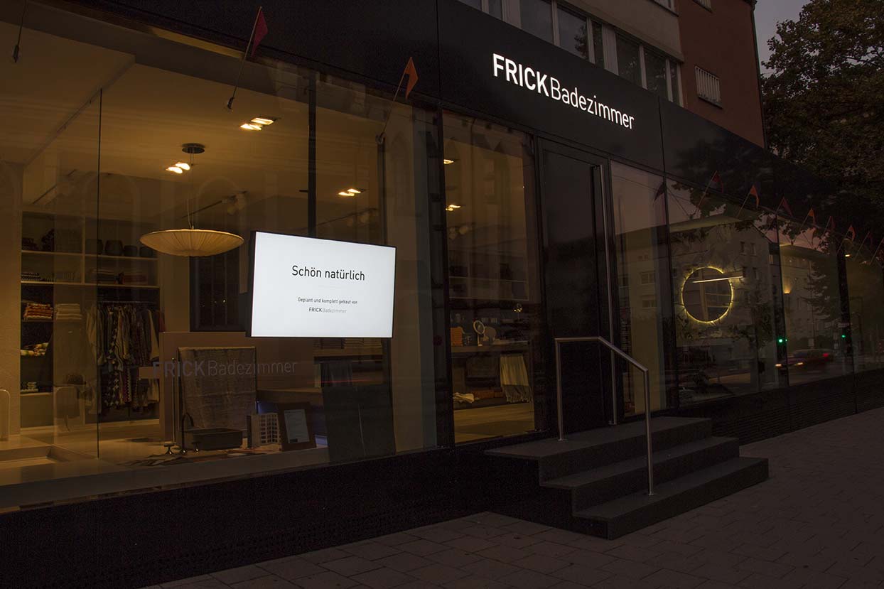 Außenansicht des Geschäfts FRICK Badezimmer bei Dämmerung mit beleuchtetem Schild und digitalem Bildschirm im Schaufenster.